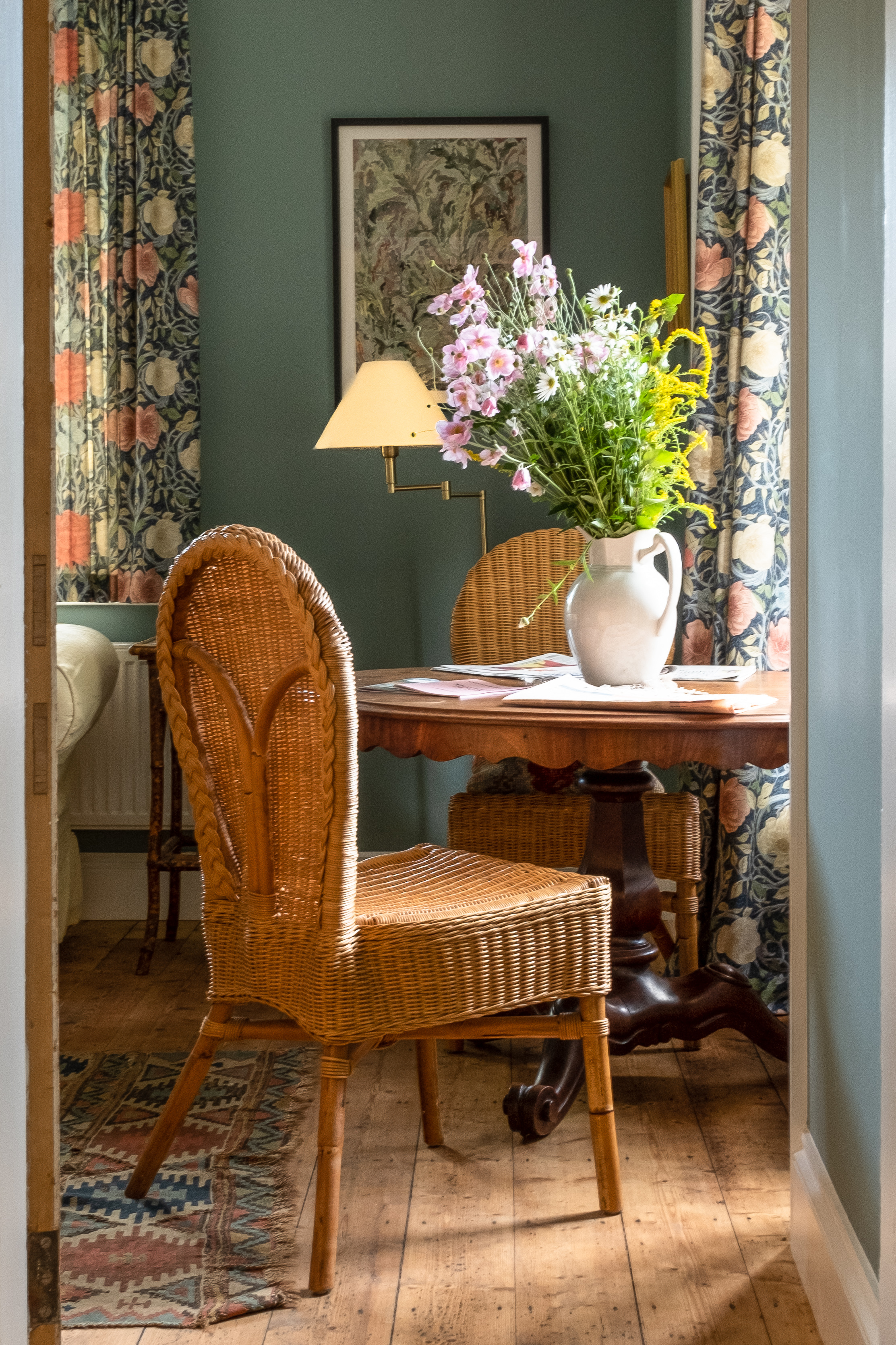 Classic wicker reading area, looking out of the window, with a beautiful bunch of flowers styled in a white, ceramic milk jug, on the table. 