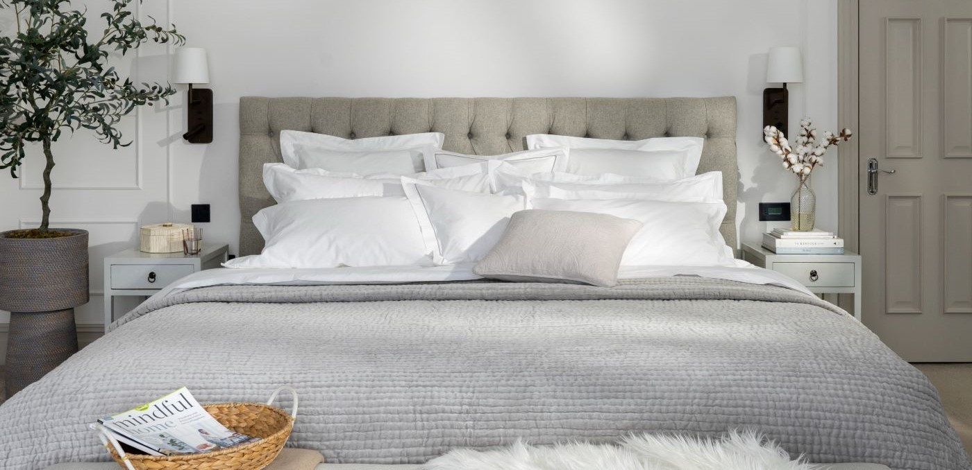 Dreamy shot of crisp 600TC White bed linen with a grey channel quilted cotton bedspread draped over the end of the bed. Dappled lighting and neutral accessories.