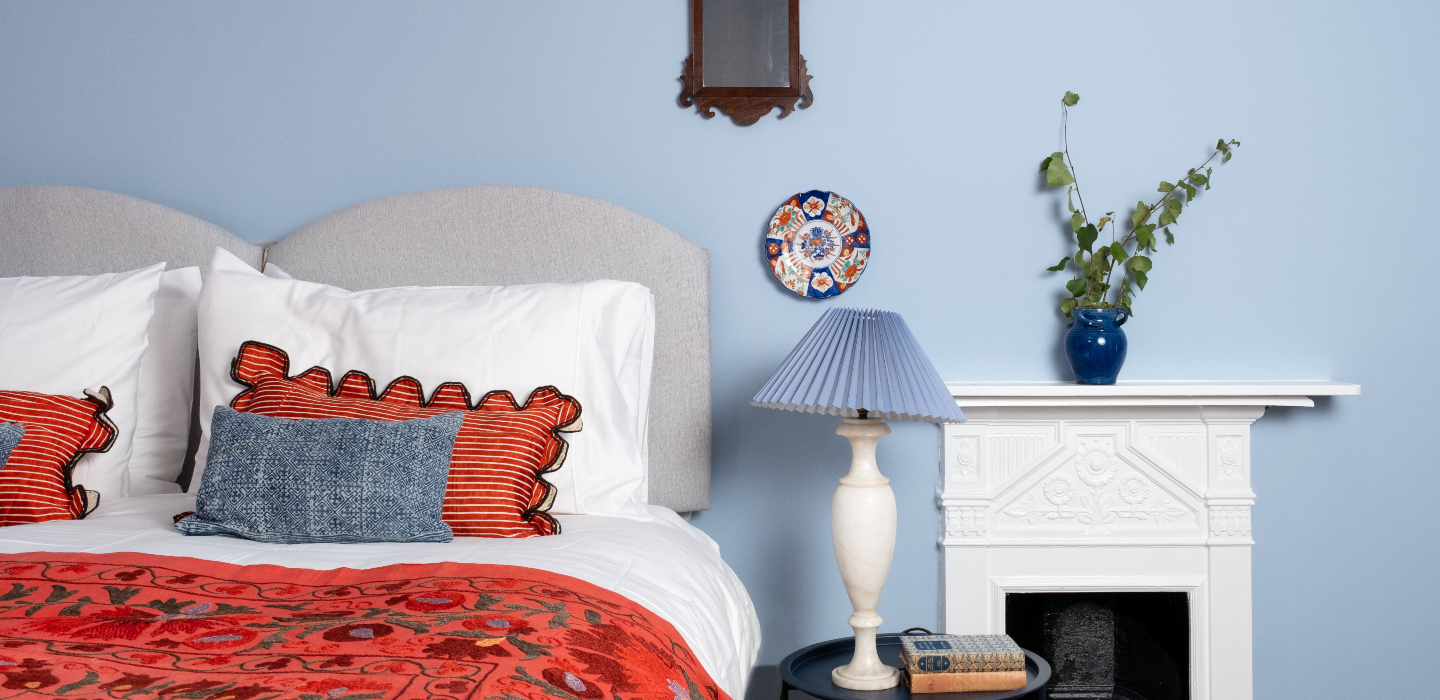 A wonderfully bright blue bedroom with crisp Soak&Sleep white bed linen and orange throws and cushions. The room has a stunning, white, classic fireplace and is styled with homely ornaments.