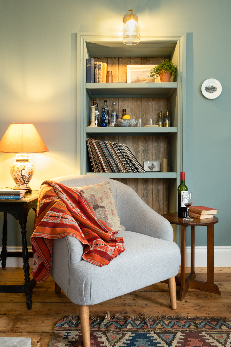 A warm, cosy shot of a reading corner in a light teal room with an arm chair styled with an orange, autumnal throw. A wooden side table holding a bottle of red wine and some books. Plus, an integrated bookshelf with some homely ornaments, in the middle.